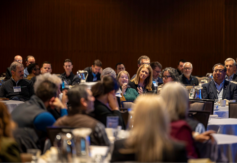 Woman speaking from sitting position with microphone at crowded event.